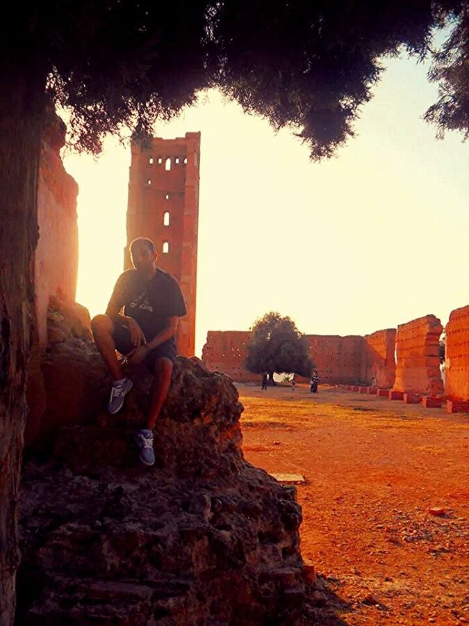 architecture, building exterior, lifestyles, built structure, leisure activity, clear sky, men, tree, photographing, sunlight, city, photography themes, sitting, casual clothing, full length, holding, outdoors, rock - object