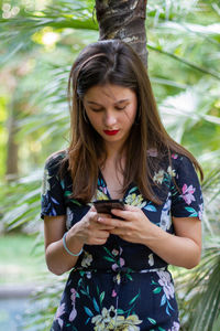 Young woman using mobile phone while standing against tree