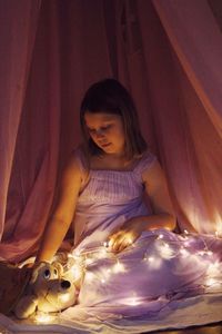Girl with stuffed toy and illuminated string lights sitting on bed
