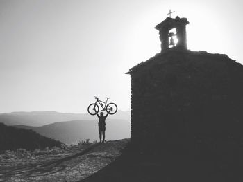 Rear view of man lifting bicycle by chapel on mountain