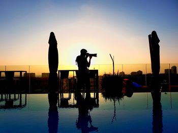 Silhouette people photographing by swimming pool against sky during sunset