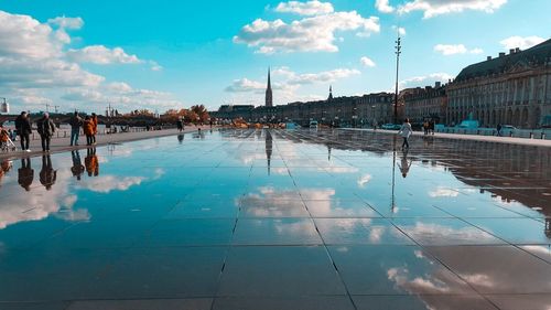 Reflection of people on wet swimming pool in city against sky