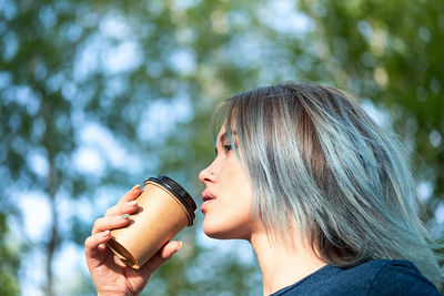 Blue haired woman drinks take away coffee outdoors on nature background.
