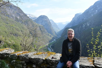 Portrait of man sitting on retaining wall against mountains