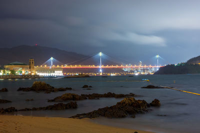 Illuminated suspension bridge over sea against cloudy sky