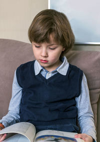 Cute boy reading book sitting on sofa at home