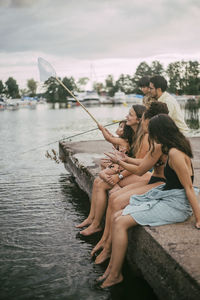 Family and friends fishing while sitting on pier by lake during summer