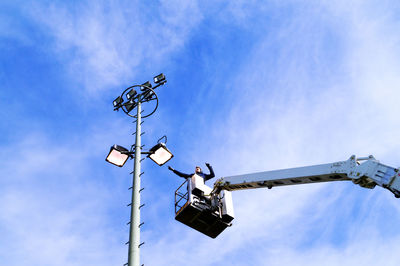 Low angle view of man standing on cherry picker by floodlight against sky