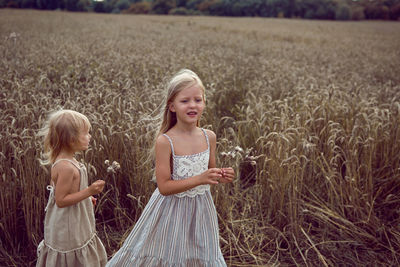 Two sisters blonde girls in dresses stand in a field with wheat ears in the summer