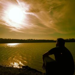 Rear view of man sitting by lake against sunset sky