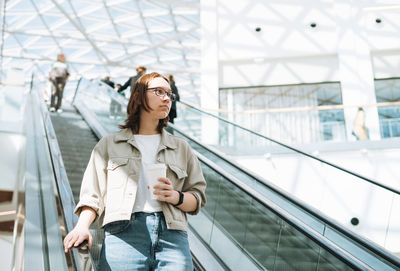 Young brunette teenager girl student in glasses on escalator in shopping mall public place