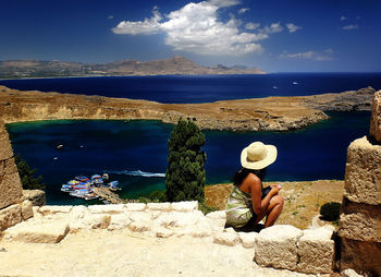 Rear view of woman sitting on rock by sea against sky