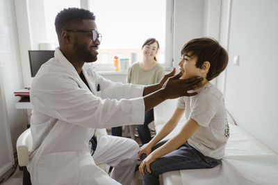 Smiling male pediatrician examining neck of boy sitting on bed at healthcare center