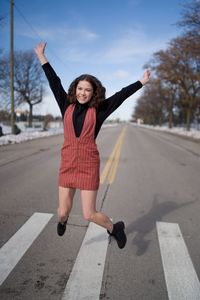 Full length portrait of a young woman with arms raised while jumping in a crosswalk