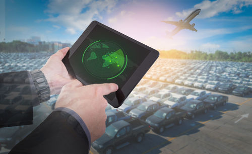 Cropped hands of man using digital tablet against airplane flying over parking lot