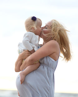 Pregnant mother kissing daughter at beach against sky