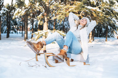 Happy woman with blonde hair in winter clothes having fun with sleigh in snowy winter forest