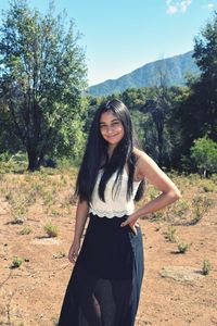 Portrait of young woman smiling while standing with hand on hip at field