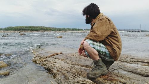 Hipster crouching on rock formation in river against sky