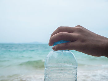 Cropped image of human hand holding bottle against sea and sky
