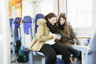 Happy young female friends using mobile phone while traveling in train