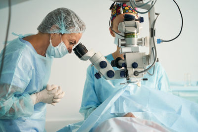 Adult female doctor in sterile mask and ornamental medical cap looking through surgical microscope against crop coworker in hospital