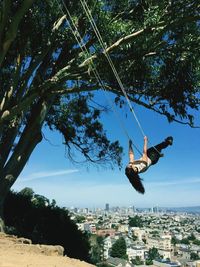 Full length of woman on swing by tree against cityscape