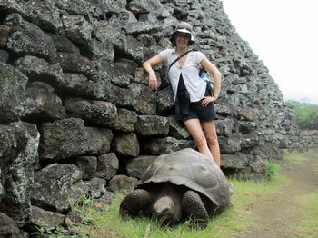 Low angle view of happy woman standing by stone wall in front of giant tortoise on field