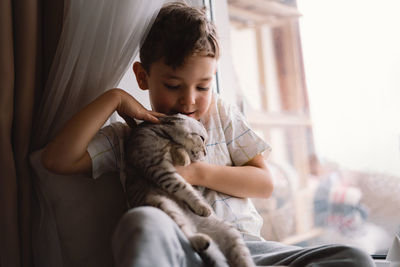 Cute boy playing with kitten sitting on the windowsill near the window. cozy home.