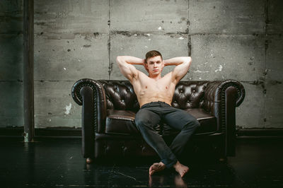 Portrait of shirtless muscular man sitting on armchair against wall