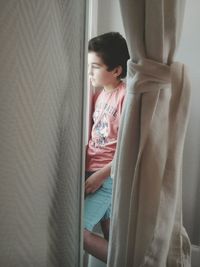 Thoughtful boy standing by window seen through curtains
