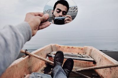 Low section of man holding mirror with reflection on boat at beach