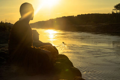 Side view of man sitting by lake against sky during sunset