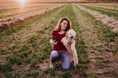 Smiling woman holding dog taking selfie on mobile phone in farm