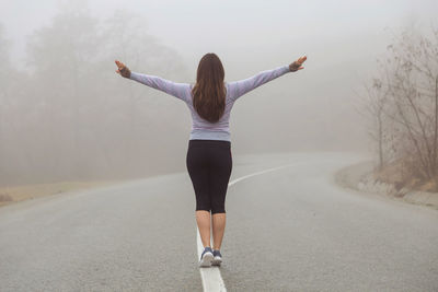 Rear view of mid adult woman with arms outstretched standing on road during foggy weather