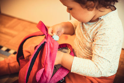 Small girl sitting on floor and packing her backpack while preparing for first day at kindergarten.