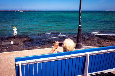 Old woman taking mobile picture of a fisherman on a bench