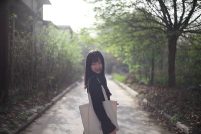 Portrait of young woman with shoulder bag standing on road amidst trees