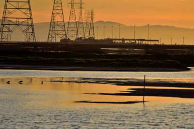 Silhouette electricity pylon by sea against sky during sunset