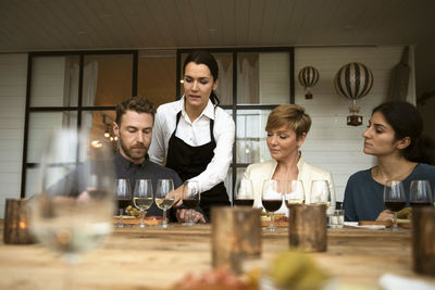 Woman wearing apron talking to business people while pointing at wineglass on table