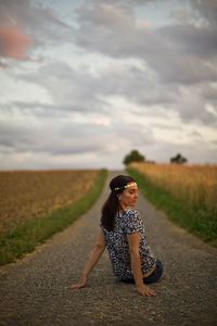 Rear view of woman sitting on road amidst field against cloudy sky