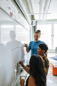 Female teacher assisting students drawing diagram on whiteboard in classroom at elementary school