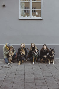 Full length portrait of smiling pet owners with dogs on footpath against wall in city