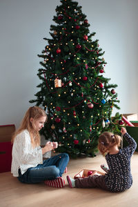 Siblings sitting by christmas tree against wall at home