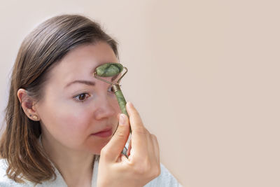 Young caucasian woman doing face massage with green jade roller on beige background