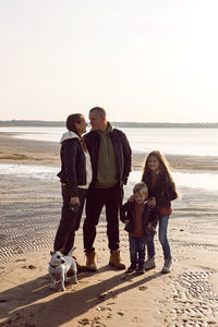 Family in a leather jacket stands along the beach with their dog in autumn