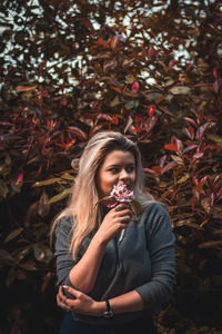 Smiling young woman with flower standing against trees 