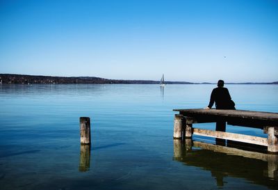 Rear view of man sitting on pier over lake against clear sky