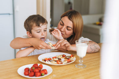 Cute toddler boy with mother having breakfast with puncakes and berries and glass of milk in kitchen 