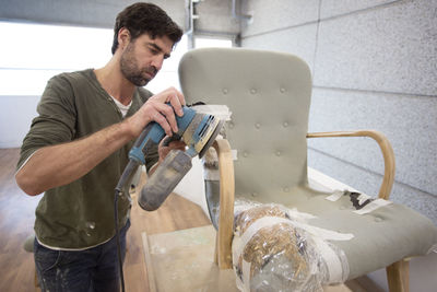 A white man power sanding the arm of an antique chair